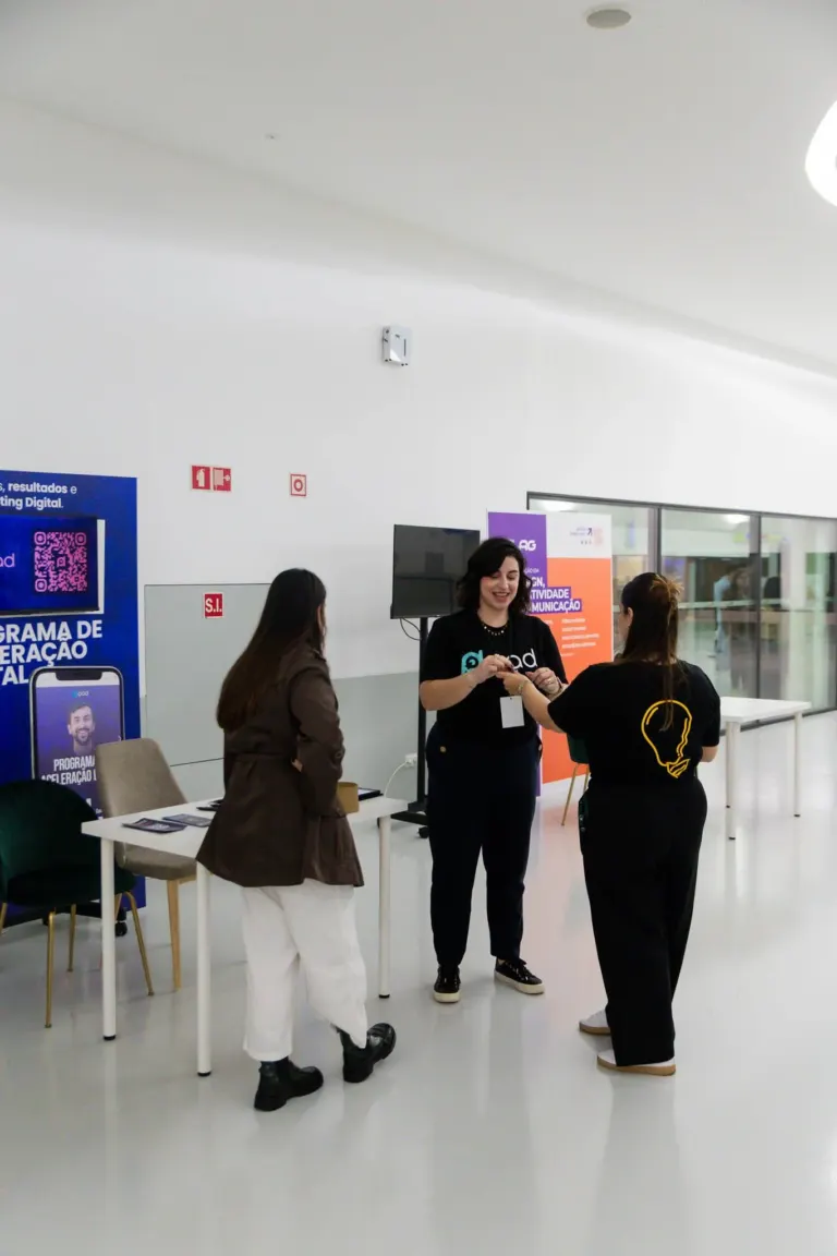 Três pessoas trocando um aperto de mão em um evento com banners ao fundo, um deles está sorrindo.