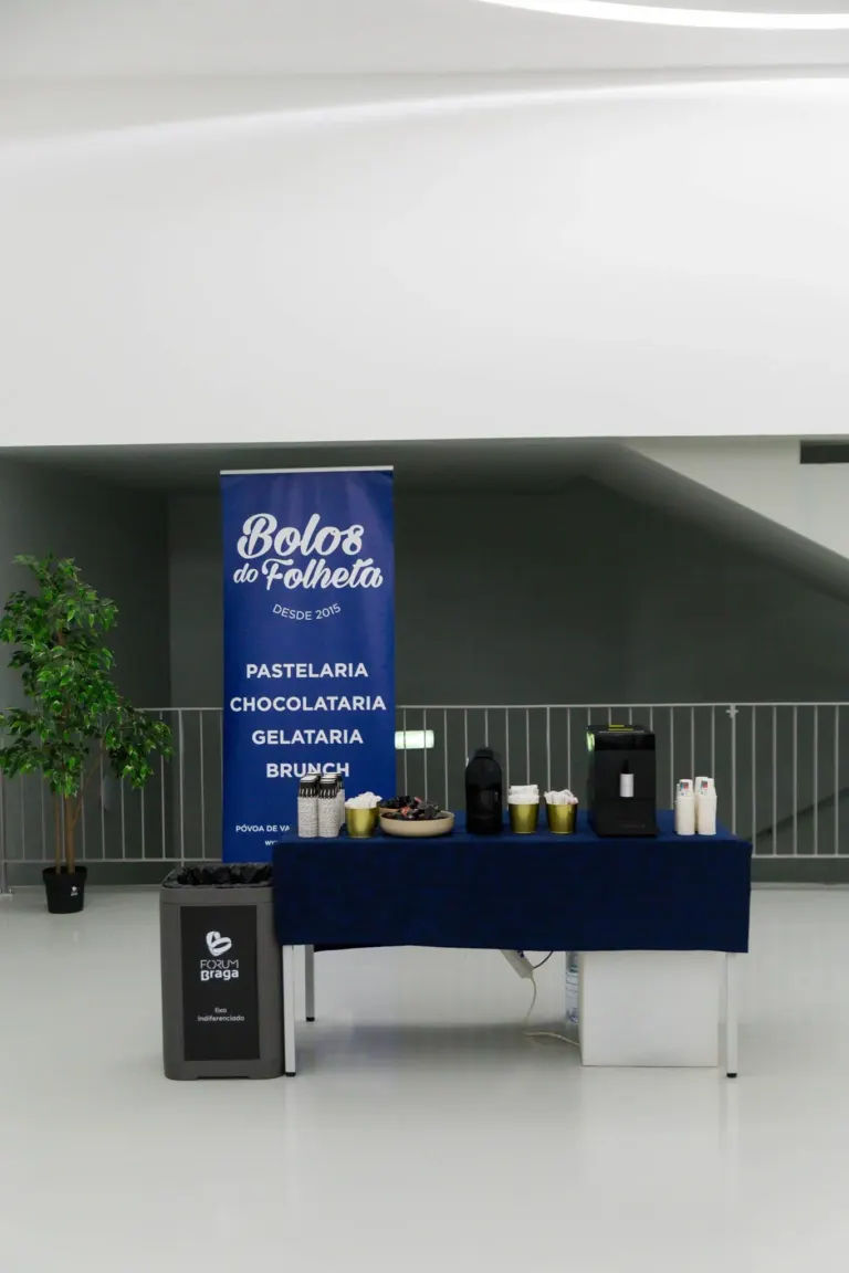 Mesa de café da manhã com máquina de café, copos, tigela com frutas, e banner azul com texto 'Bolos do Folheta, desde...
