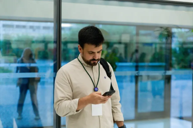 Homem com camiseta bege, mochila e crachá usando celular em frente a uma entrada de vidro, com uma mulher ao fundo caminhando