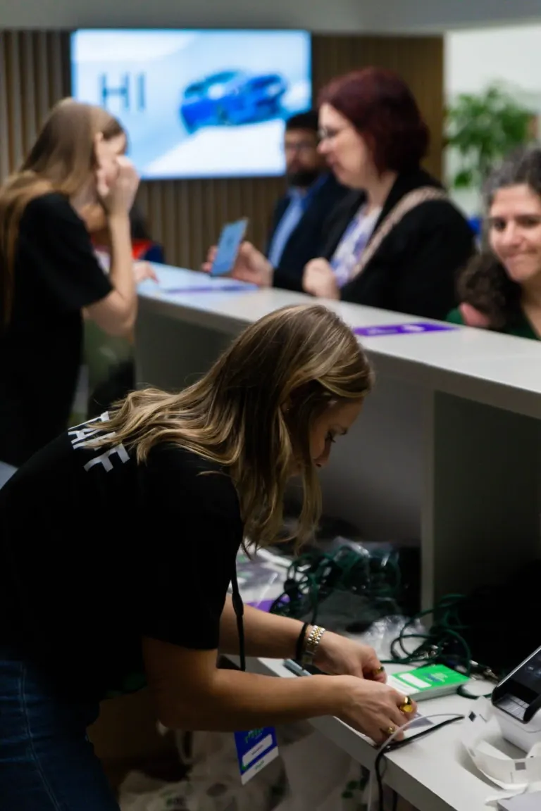 Mulher ajustando equipamentos eletrônicos em uma mesa, com várias pessoas conversando ao fundo