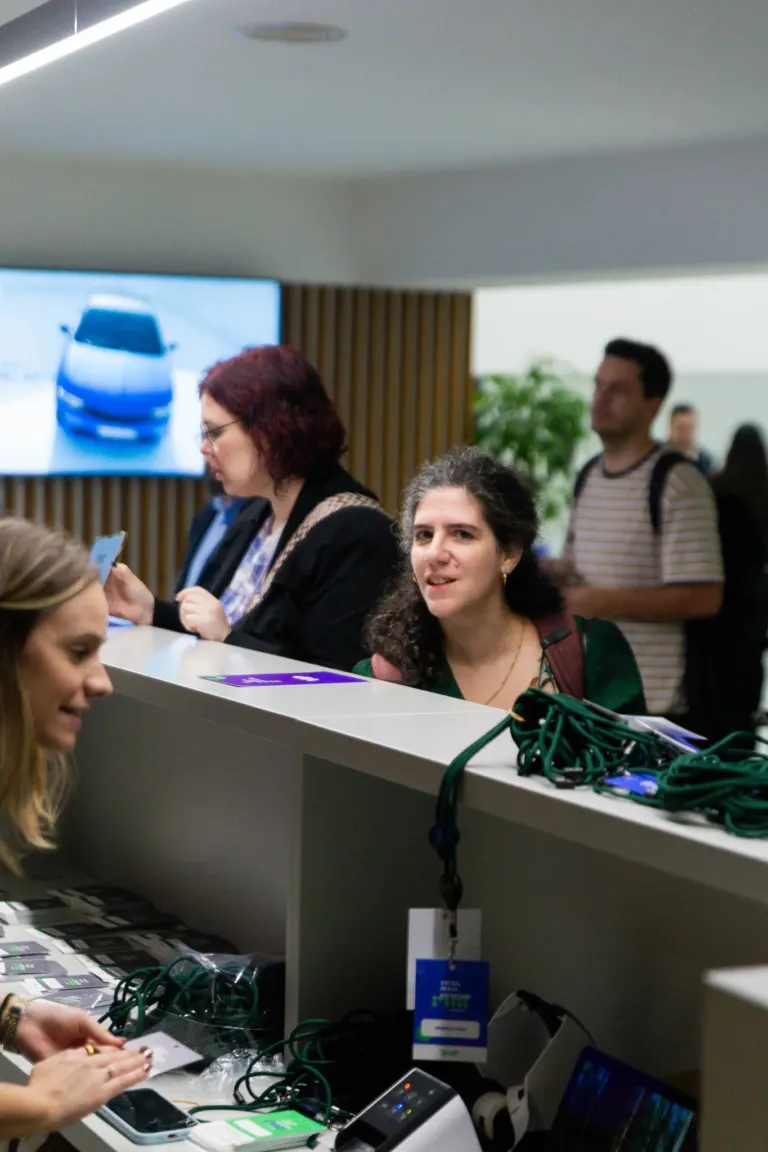 Pessoas na recepção de um evento, com cords e crachás, uma mulher com cabelos cacheados escuros conversa com alguém...
