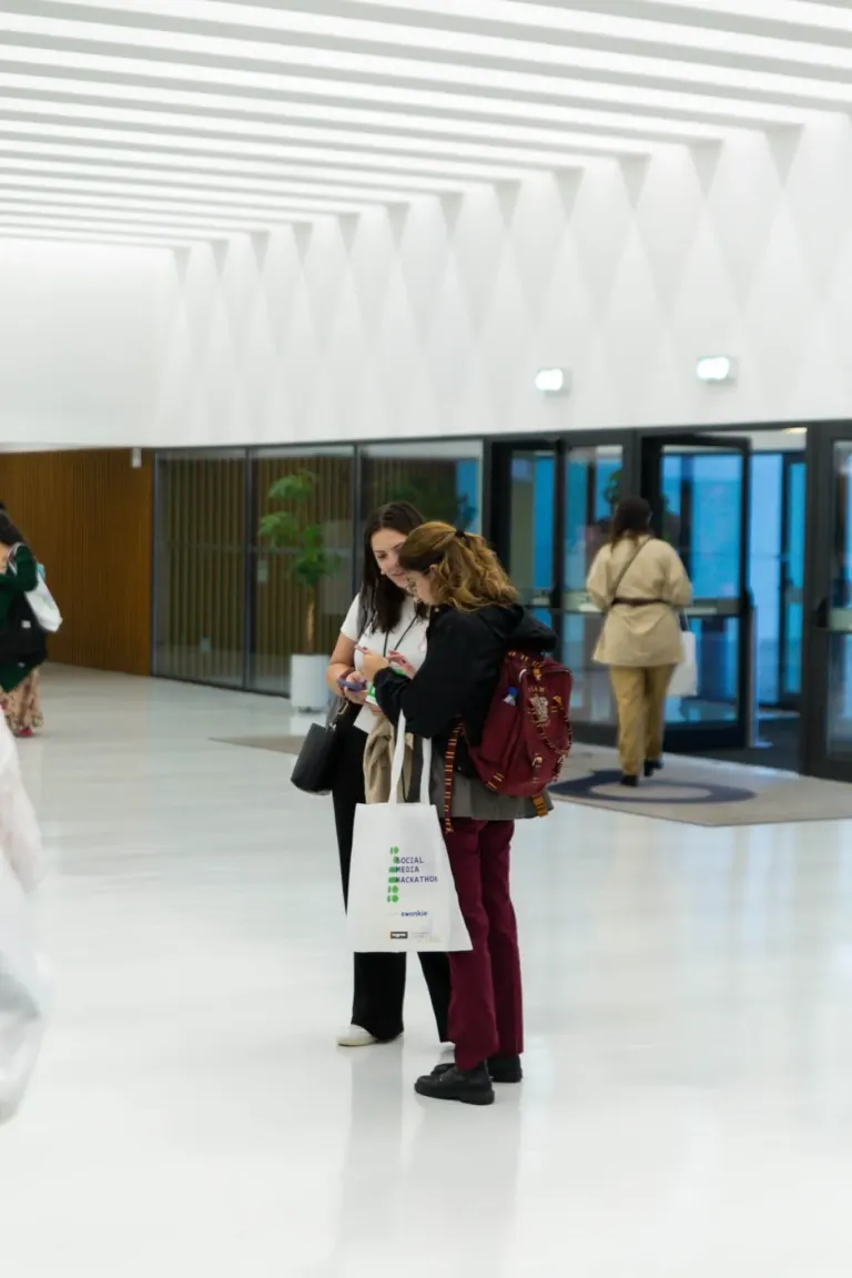 Duas pessoas olhando para um celular em uma entrada de aeroporto, com outras pessoas ao fundo e portas de vidro.