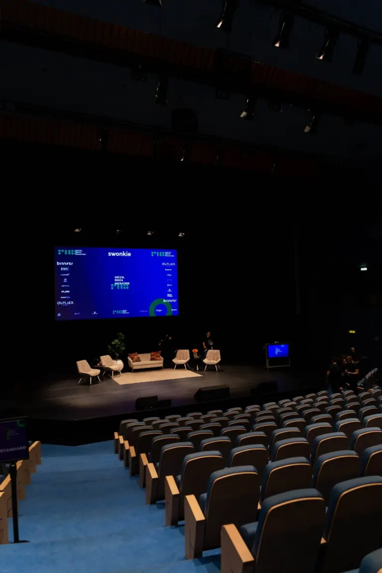 Auditório vazio com cadeiras azuis, palco com poltronas, televisão e tela grande exibindo o texto "Social Media Marathon"...