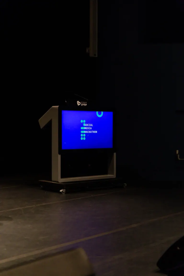 Tela de televisão exibindo o texto 'SOCIAL MEDIA HACKATHON' em fundo azul, posicionada em um suporte móvel em um ambiente...