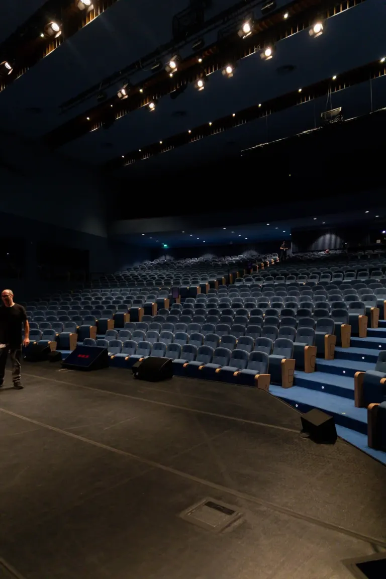 Auditório vazio com cadeiras azuis, palco com equipamentos de som, e pouca iluminação.