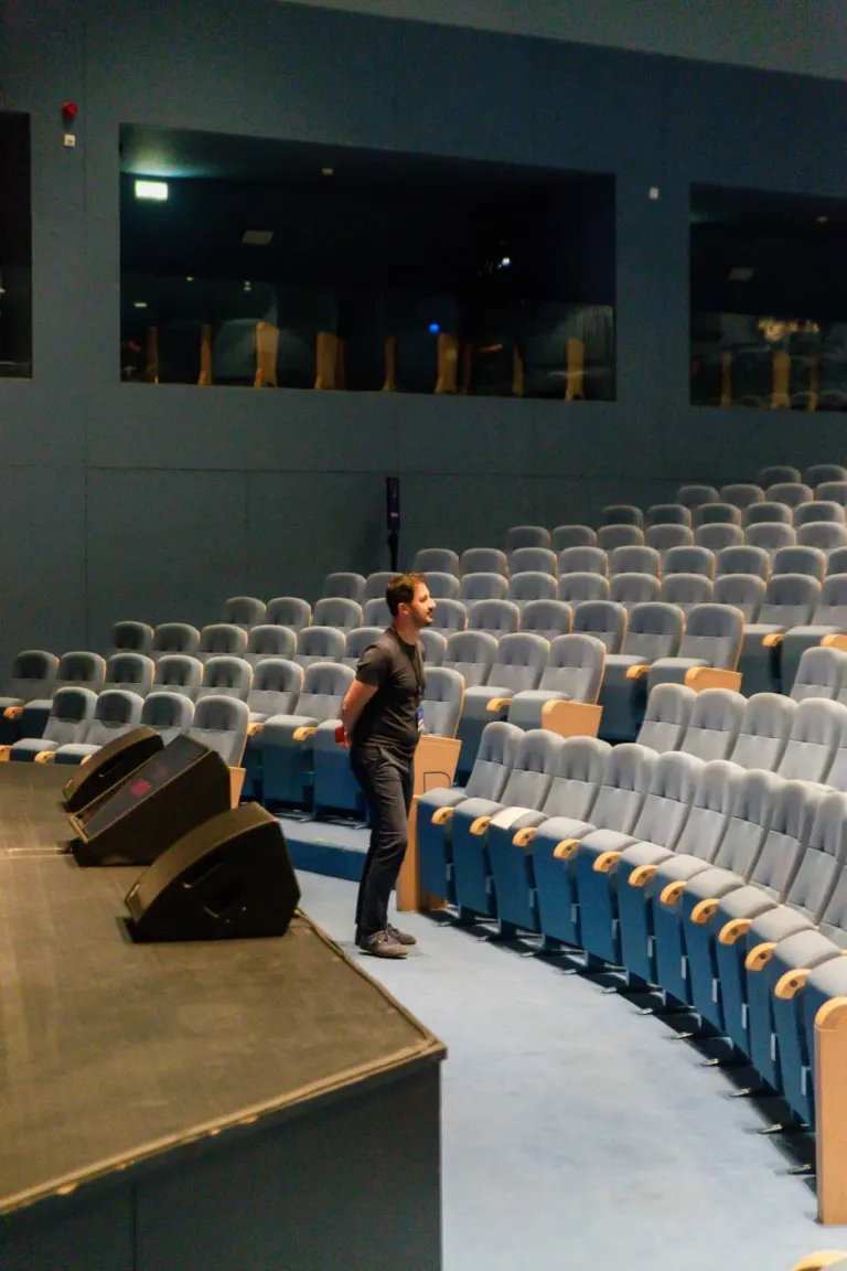 Homem de terno escuro caminhando em uma sala de auditório vazia com cadeiras azuis e palco com equipamento de som.