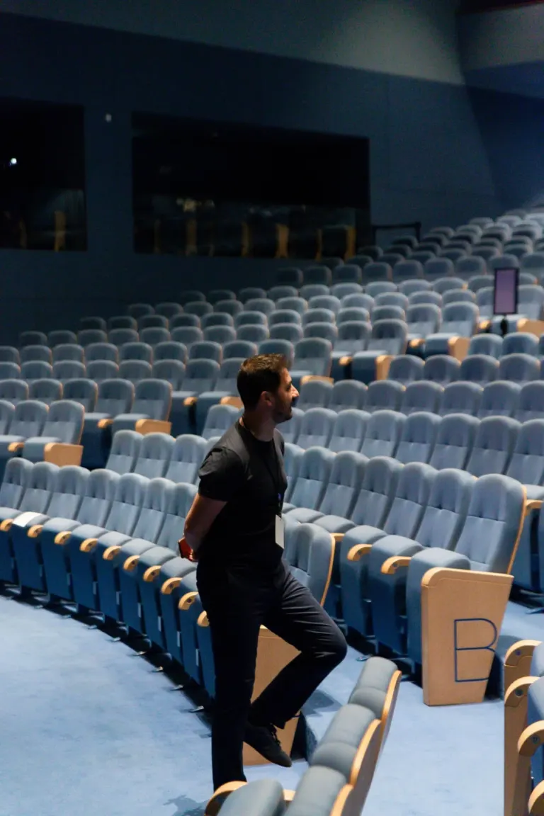 Homem de terno escuro caminhando entre assentos de teatro ou auditório com assentos azuis.