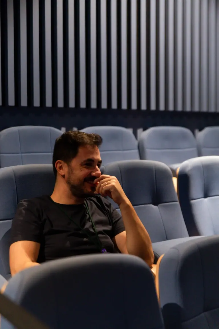 Homem sorridente sentado em uma sala de cinema com cadeiras azuis, de fundo uma parede com painéis verticais escuros.