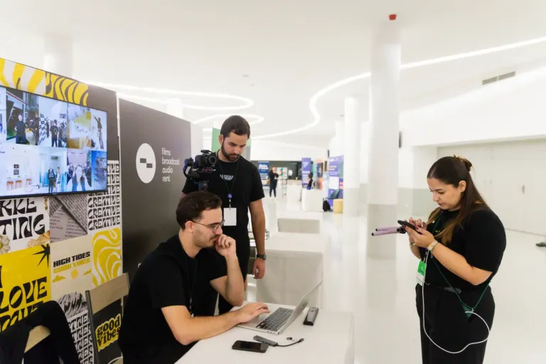 Três pessoas em uma sala de convenções, uma sentada usando um laptop com um smartphone ao lado, uma de pé com uma câmera...