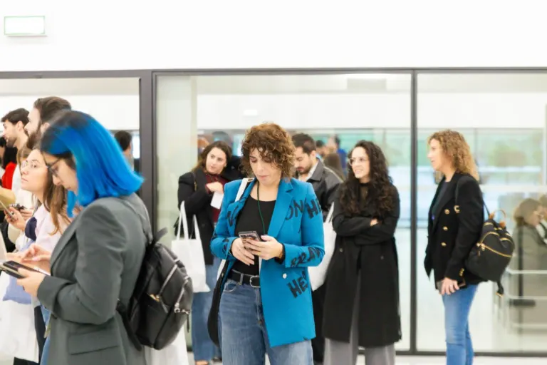Várias pessoas esperando em uma fila, algumas olhando seus celulares, dentro de um ambiente provavelmente de aeroporto ou...
