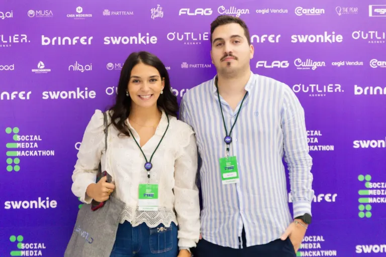 Homens e mulher posando para uma foto em evento, ao fundo há um painel roxo com logos de patrocinadores e o nome do...