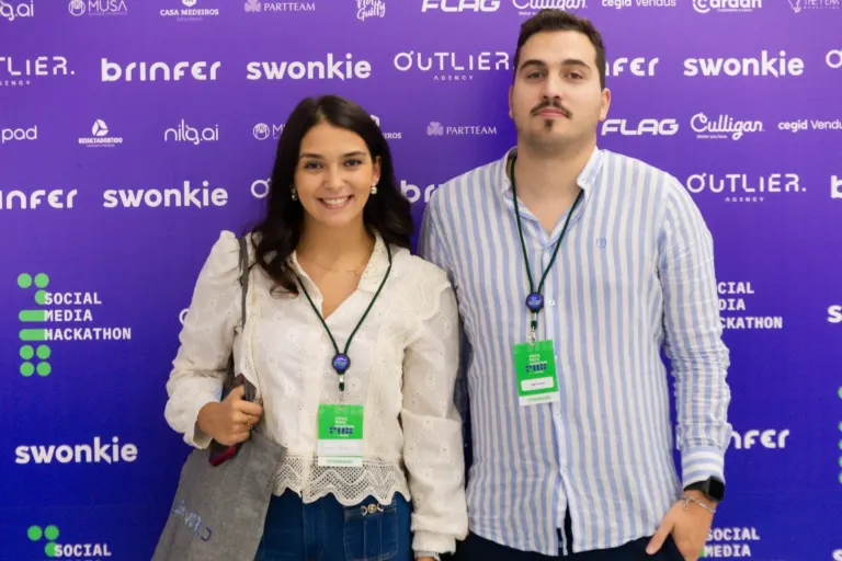 Homem e mulher sorrindo em evento, com fundo roxo contendo logos de patrocinadores e palavras 'Social Media Hackathon'.