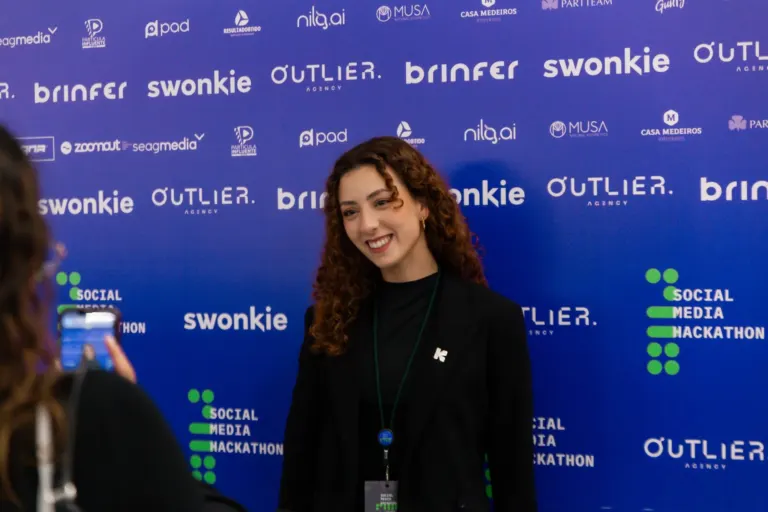 Mulher com cabelos cacheados sorrindo em evento, usando blusa preta e coroa de flores no cabelo, com fundo azul com logos...