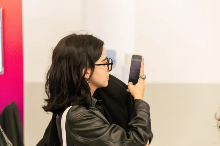 Mulher de cabelos escuros usando óculos e jaqueta preta, sentada e olhando para um celular, com uma mochila branca ao lado.
