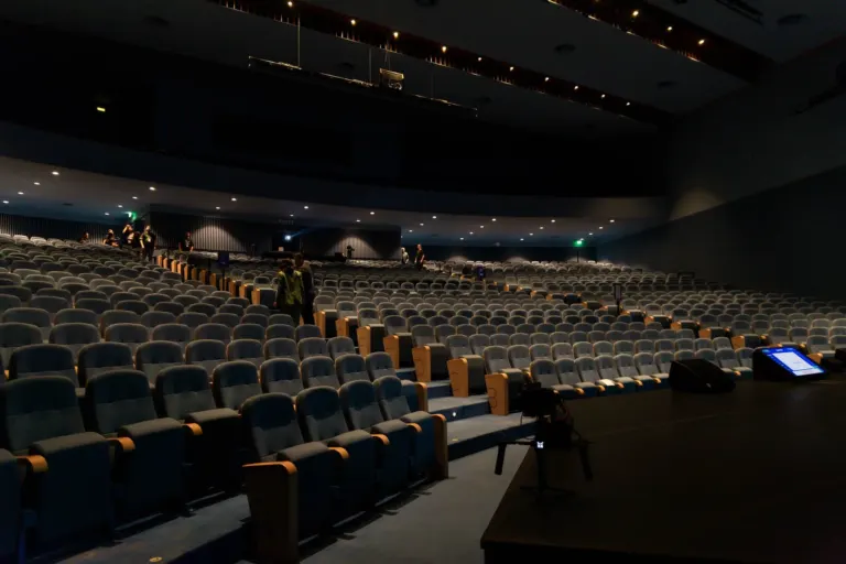 Auditório vazio com cadeiras azuis, algumas pessoas e equipamento de áudio e vídeo ao palco.
