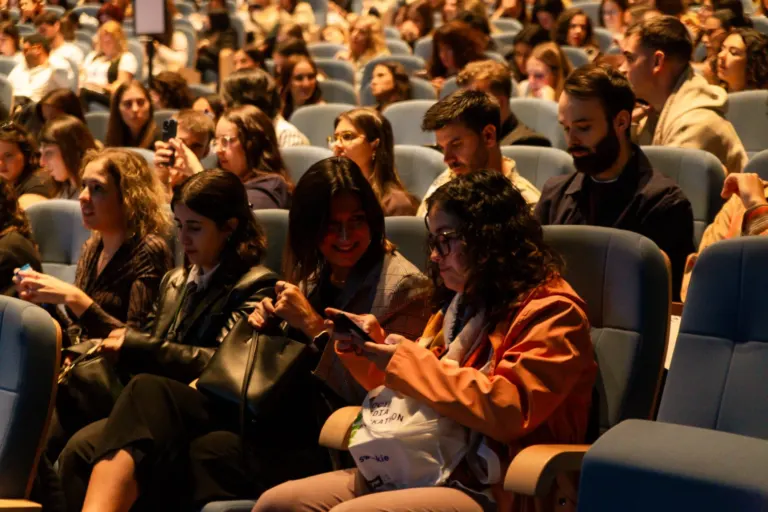 Pessoas sentadas em assentos de teatro ou auditório, participando de um evento, muitas usando roupas casuais e alguns...