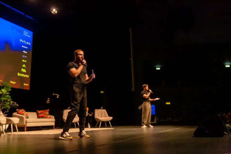 Homem com microfone no palco, vestido com camiseta preta, calça preta e tênis colorido, ao lado de outro homem também com...