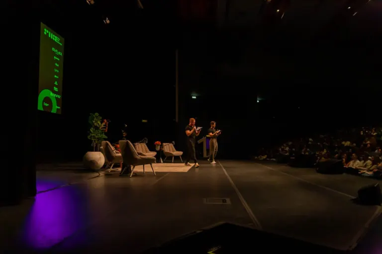Cena de uma conferência ou evento em um teatro escuro, com duas pessoas de pé na frente do público, uma lendo de um...