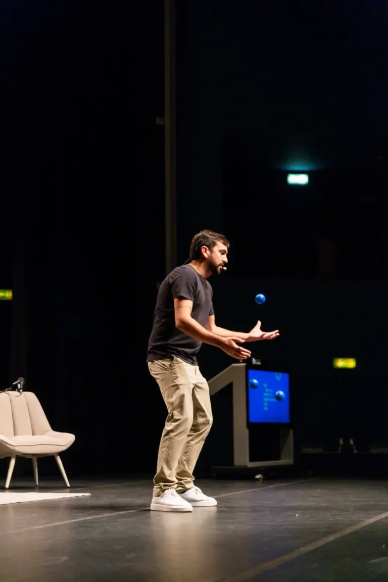 Homem de camiseta preta e calça bege, usando microfone na cabeça, participando de uma apresentação ou palestra em palco...