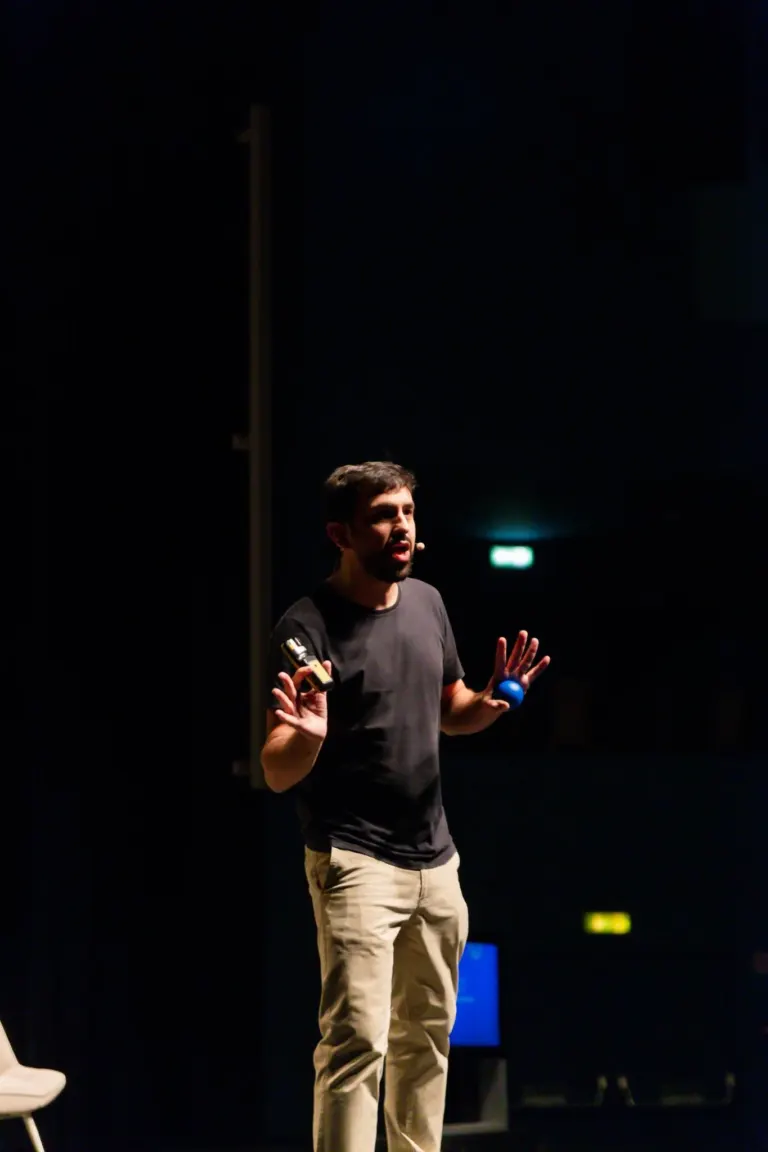 Homem de cabelo escuro e barba, usando camiseta cinza e calças bege, falando em um microfone em um palco com fundo preto.