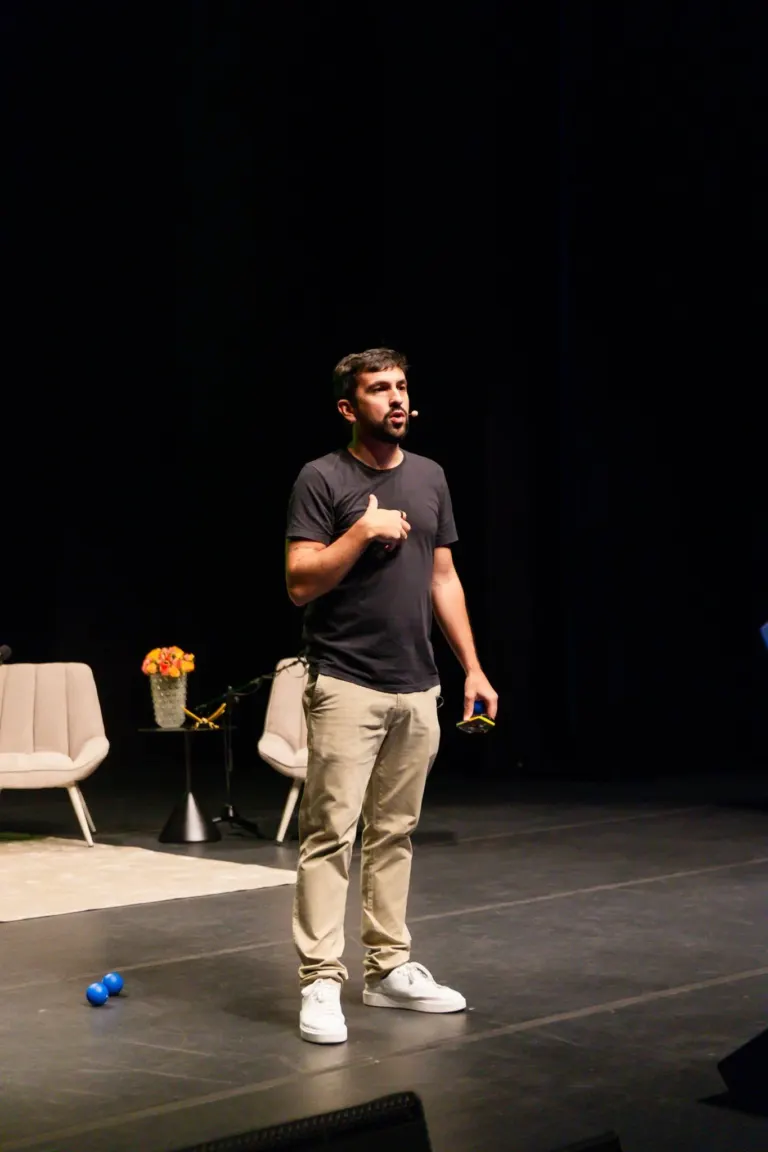 Homem de barba, com microfone na orelha, usando camiseta preta e calça bege, fala em palco escuro ao fundo, com duas...