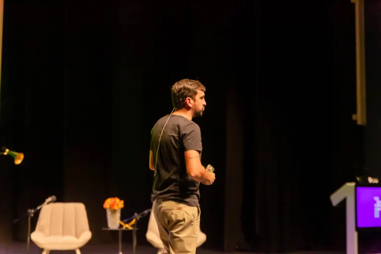 Homem com cabelo castanho curto e barba no palco, segurando controle remoto. Fundo escuro com algumas cadeiras brancas,...