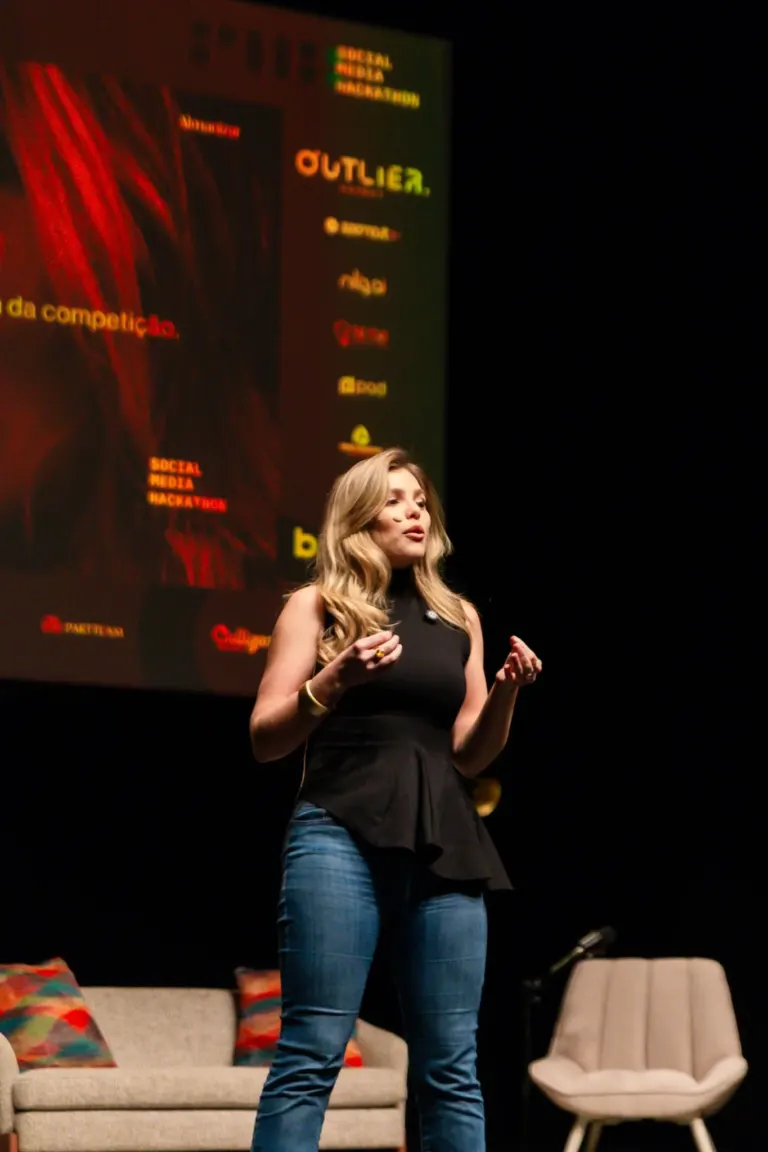 Mulher jovem falando em palco com uma tela ao fundo, decorada com almofadas coloridas em um sofá e uma cadeira branca ao...