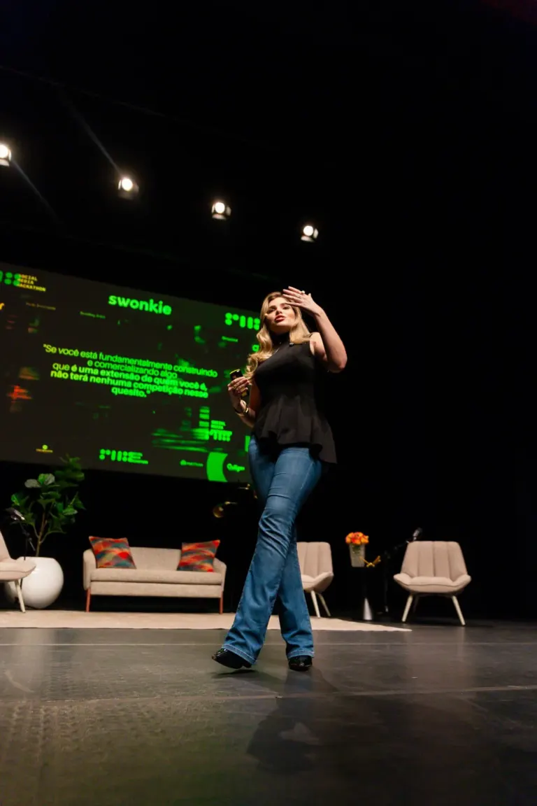 Mulher fazendo apresentação em palco, usando roupas jeans e blusa preta, ao fundo há cadeiras, uma mesa com flores, um...