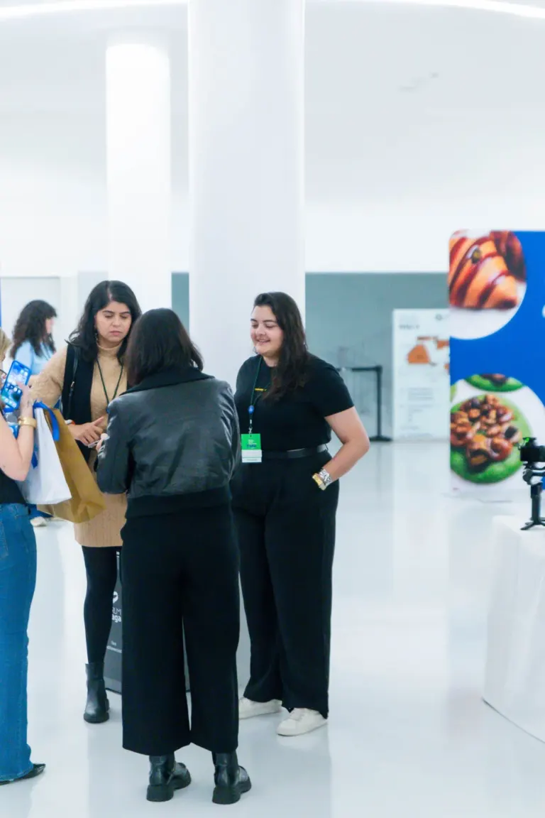 Grupo de pessoas conversando em uma feira ou exposição, com fundo branco e algumas placas de produtos ao lado.
