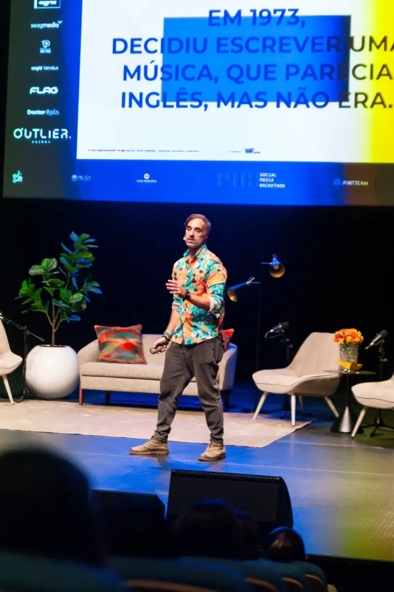 Homem de pé em palco de frente, com roupas casuais, falando para audiência durante apresentação. Fundo com sofá,...