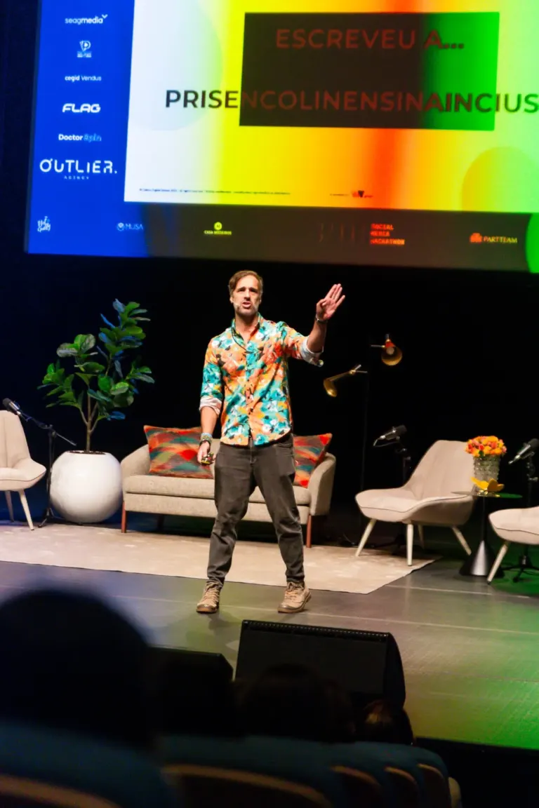 Homem de cabelo castanho, vestindo uma camisa com estampa colorida, calças cinza e botas, está apresentando em um palco...