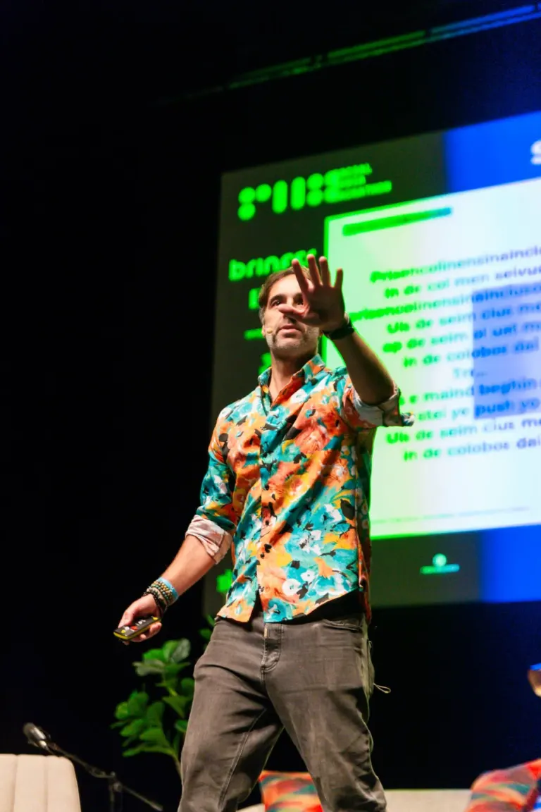 Homem de camiseta com estampa colorida e calça cinza, segurando controle remoto, durante apresentação com slide projetado...