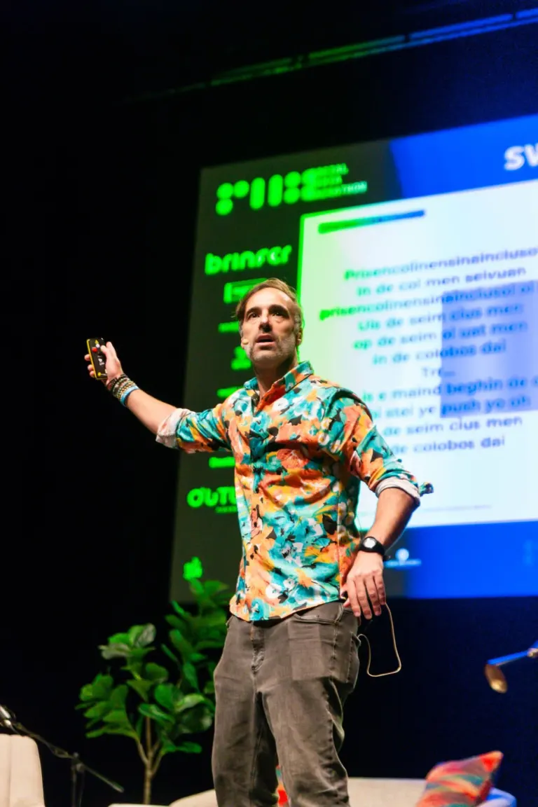 Homem de camisa floral e calça cinza, segurando um controle remoto, fazendo apresentação em palco com um projeção ao...