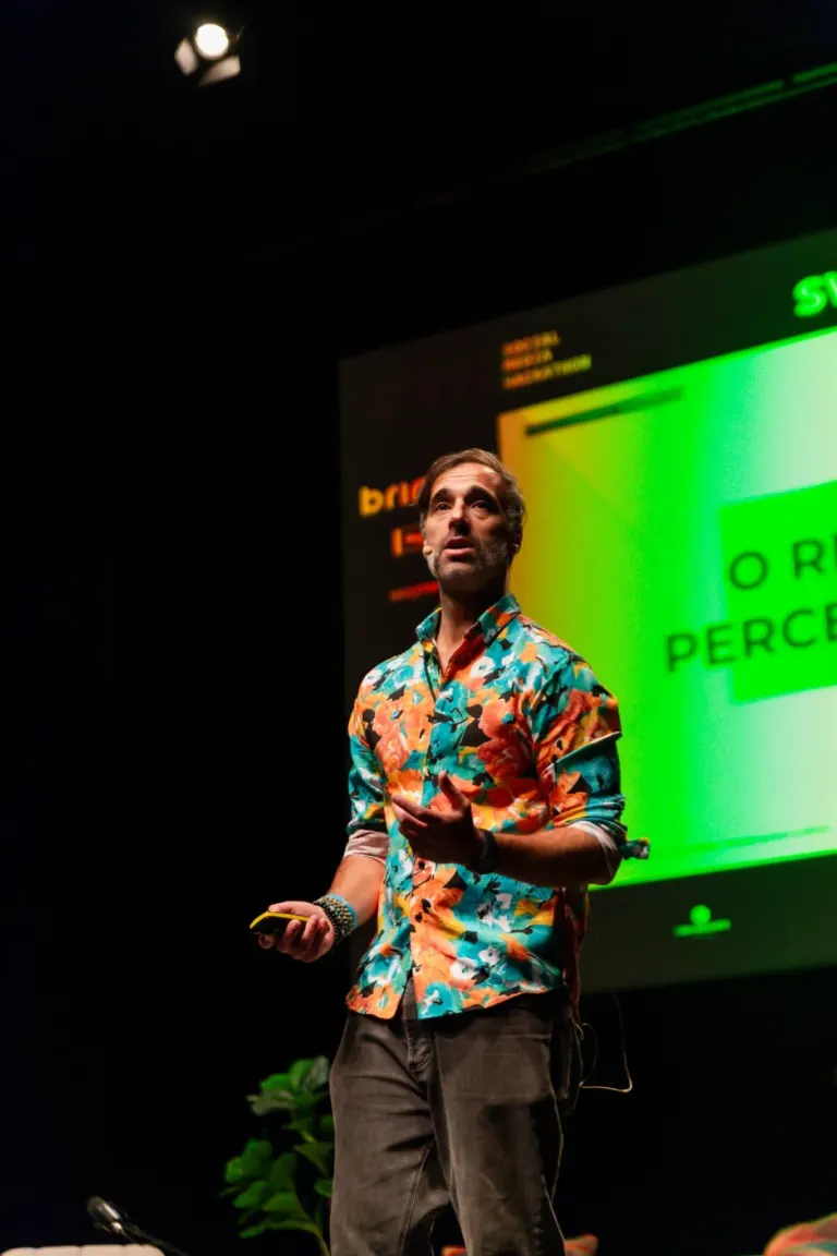 Homem falando em palco durante apresentação, usando camisa florida e segurando um celular, com tela digital visível ao fundo.