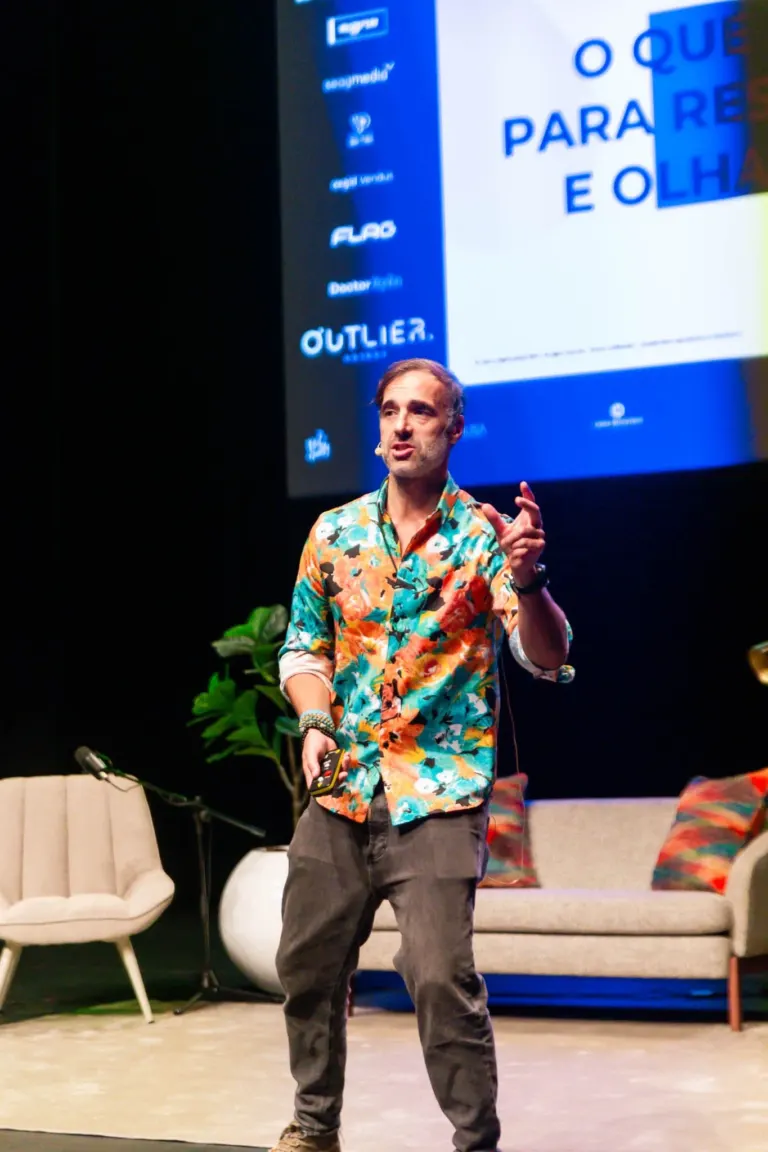 Homem falando em palco, vestindo camiseta florida, calças cinza, usando um microfone e assistido por cadeira elegante e...