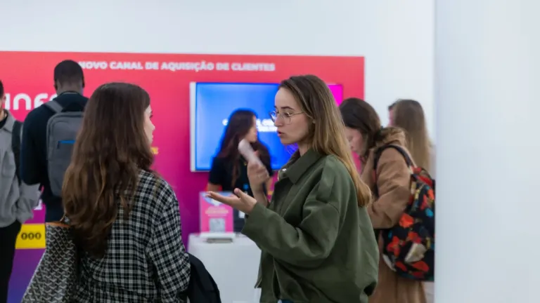 Pessoas conversando em uma feira ou evento, com fundo de painel rosa com informações em português.