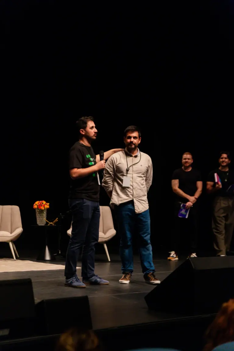 Homens no palco durante evento ou apresentação, com cadeiras ao fundo e um microfone na mão de um deles.