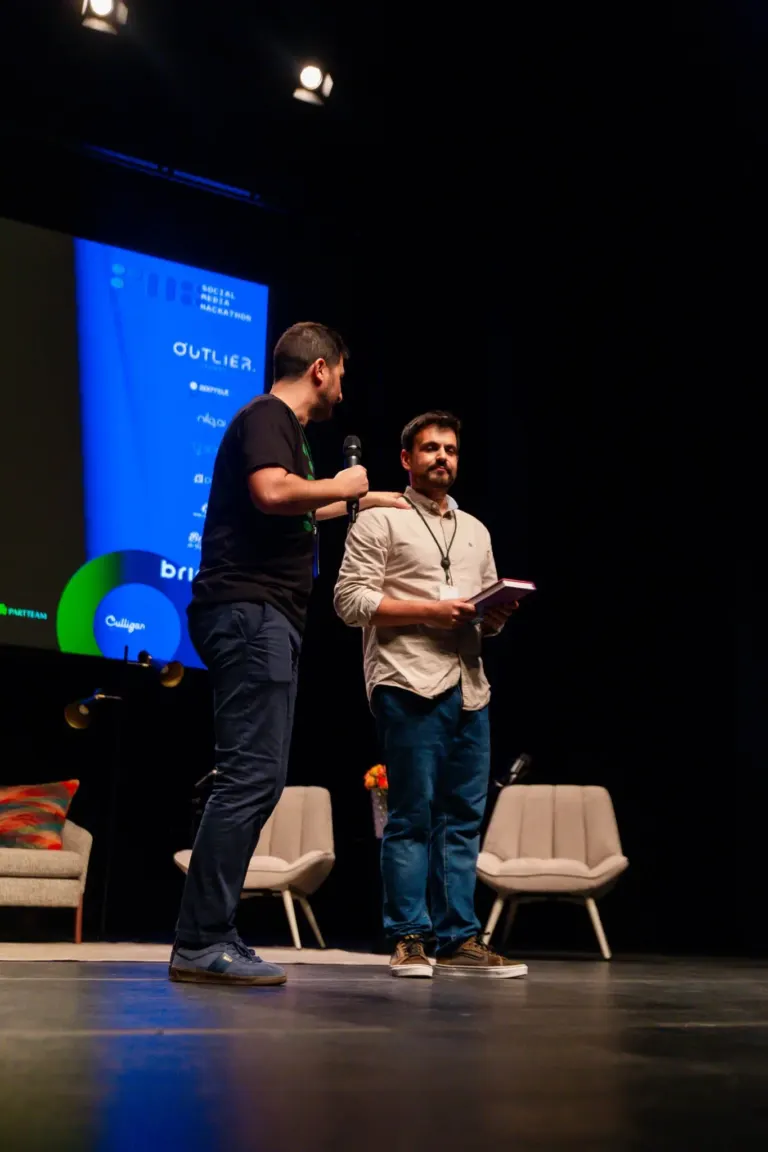 Dois homens em um palco, um segurando um microfone e o outro segurando um caderno, durante uma apresentação ou debate,...