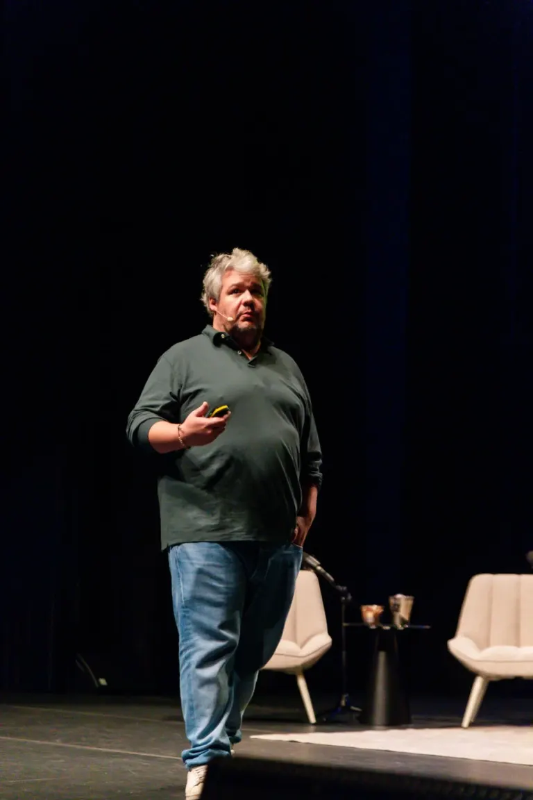 Homem com cabelo grisalho de camiseta escura e jeans azul claro, falando em palco com microfone, em um ambiente de...