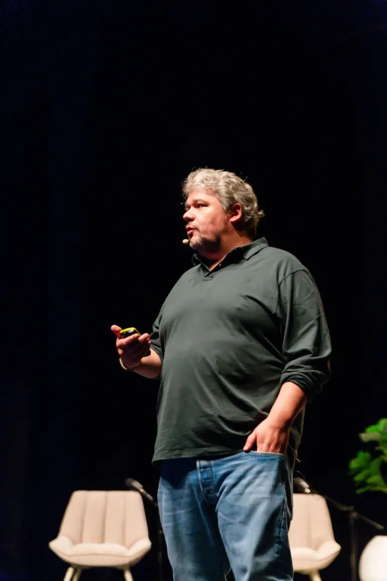 Homem com cabelos grisalhos vestindo camisa cinza escuro e jeans, segurando um telefone, de pé em um palco com fundo...