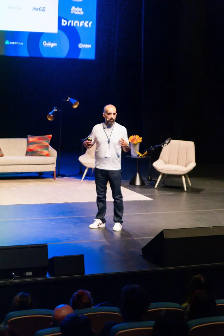 Homem com roupas casuais e microfone no palco, com móveis decorativos e tela de apresentação ao fundo, durante um evento...