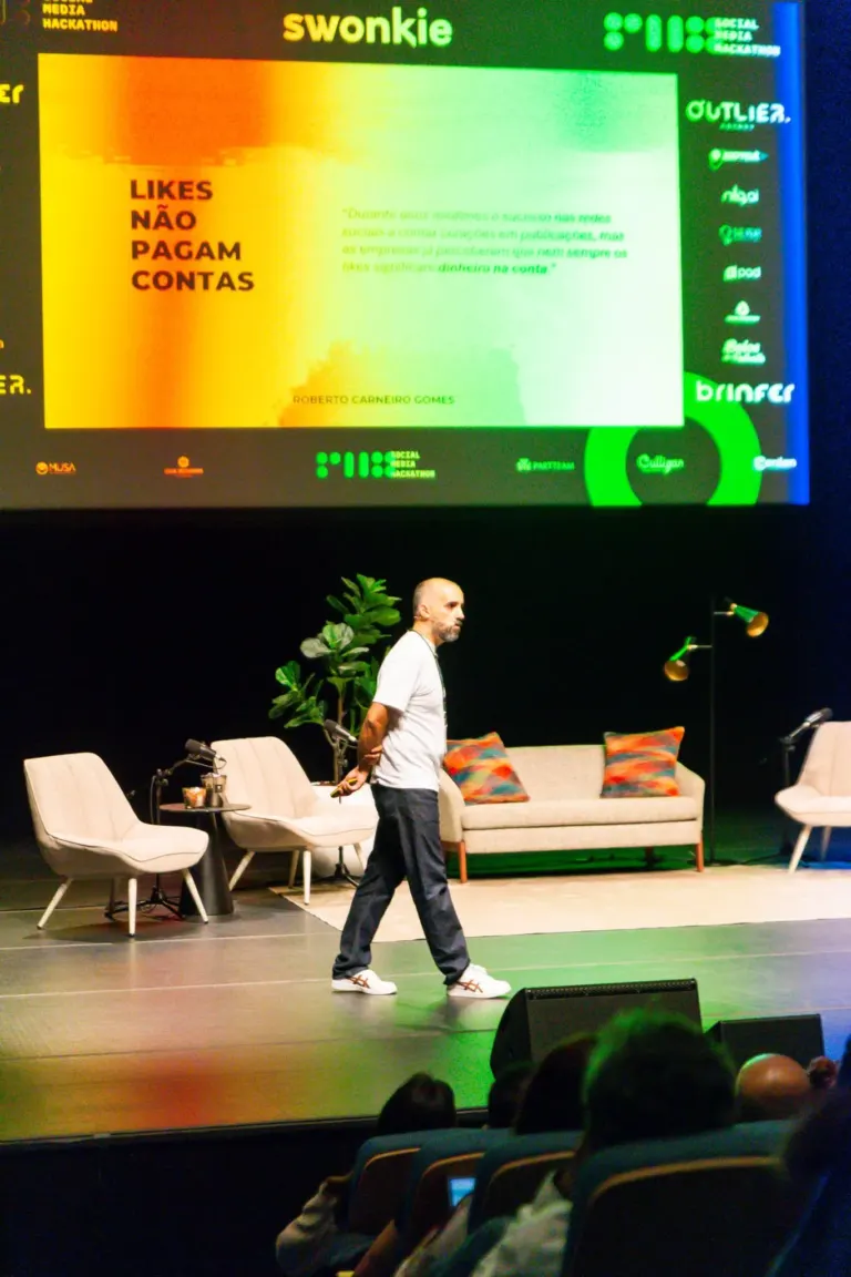 Homem de camiseta branca e calça azul caminha pelo palco de um evento de mídia social, com cadeiras e sofá ao fundo, uma...