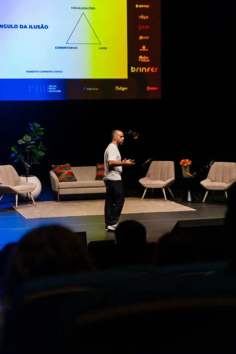 Homem em um palco apresentando diante de um tela grande com informações sobre ilusão, incluindo um triângulo com os...