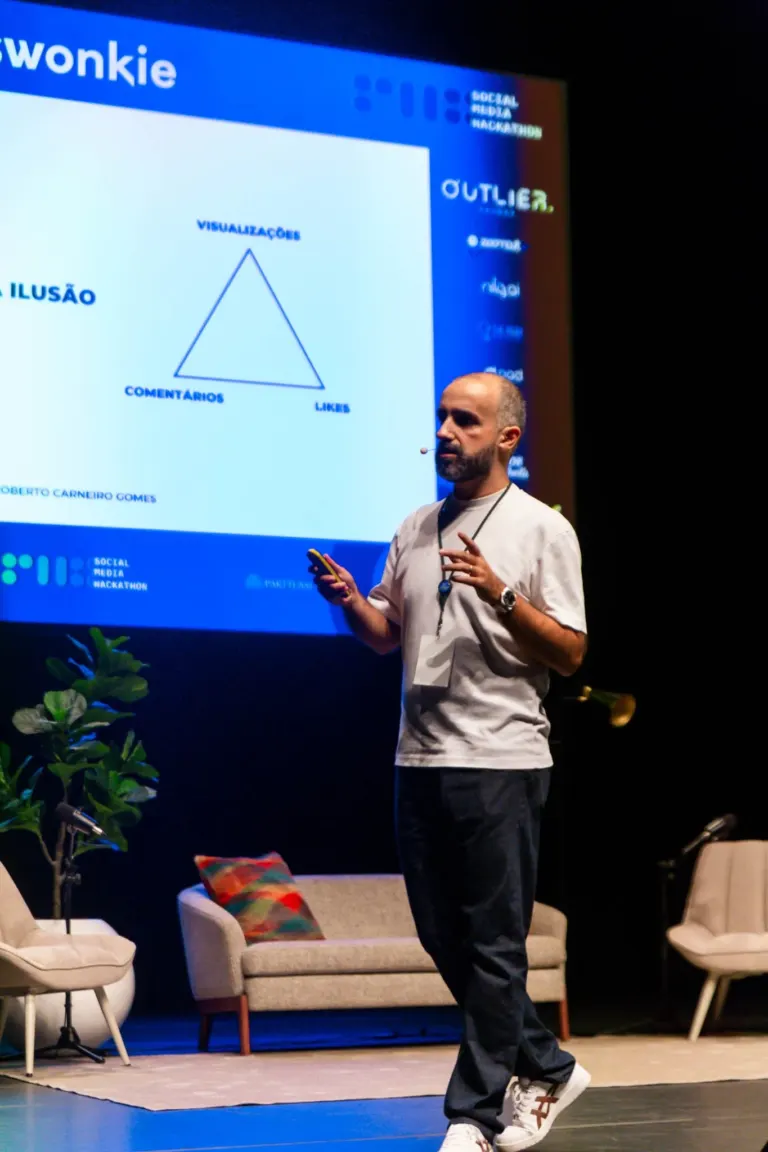 Homem de cabelo curto, barba, vestindo camiseta branca, calça escura e tênis branco, segurando um celular e gesticulando...