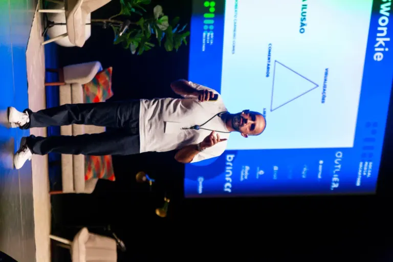 Homem apresentando em palco com microfone, ao lado de uma tela de fundo azul com texto e gráfico triangular, em um evento...