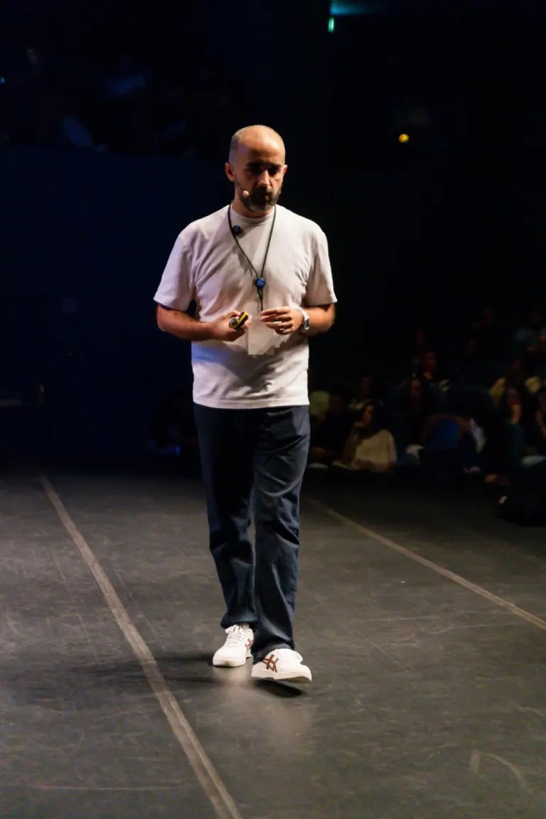Homem de terno casual, com microfone na cabeça, caminhando em palco escuro durante apresentação ou palestra.