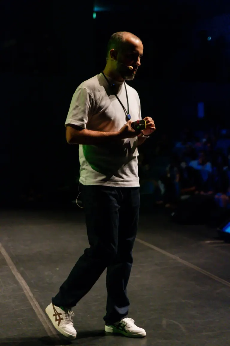 Homem em palco escuro, usando camiseta branca, calça preta, tênis branco com detalhes marrom e preto, com microfone na...
