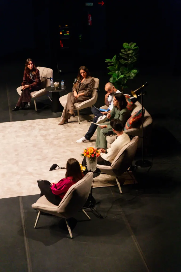 Quatro pessoas participam de um painel de discussão em um palco com cadeiras confortáveis. Há duas mulheres sentadas na...