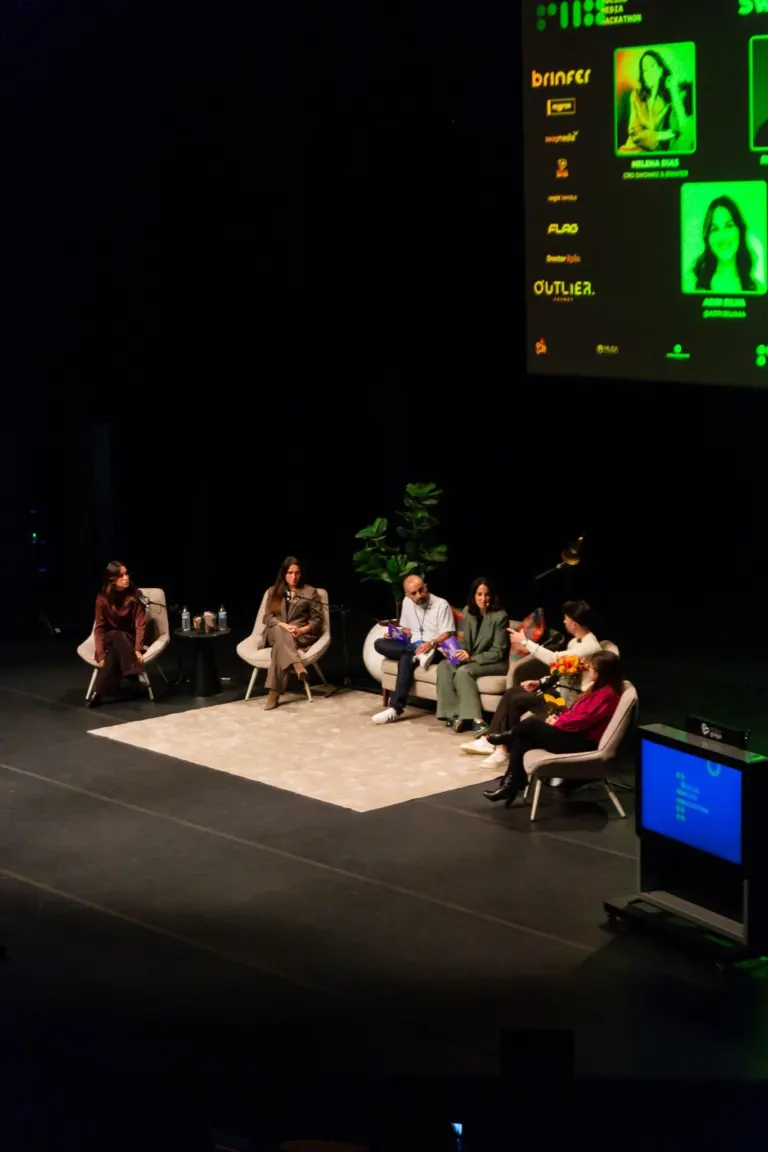 Imagem de uma conferência ou painel de discussão com várias pessoas sentadas em poltronas no palco, algumas com microfone...