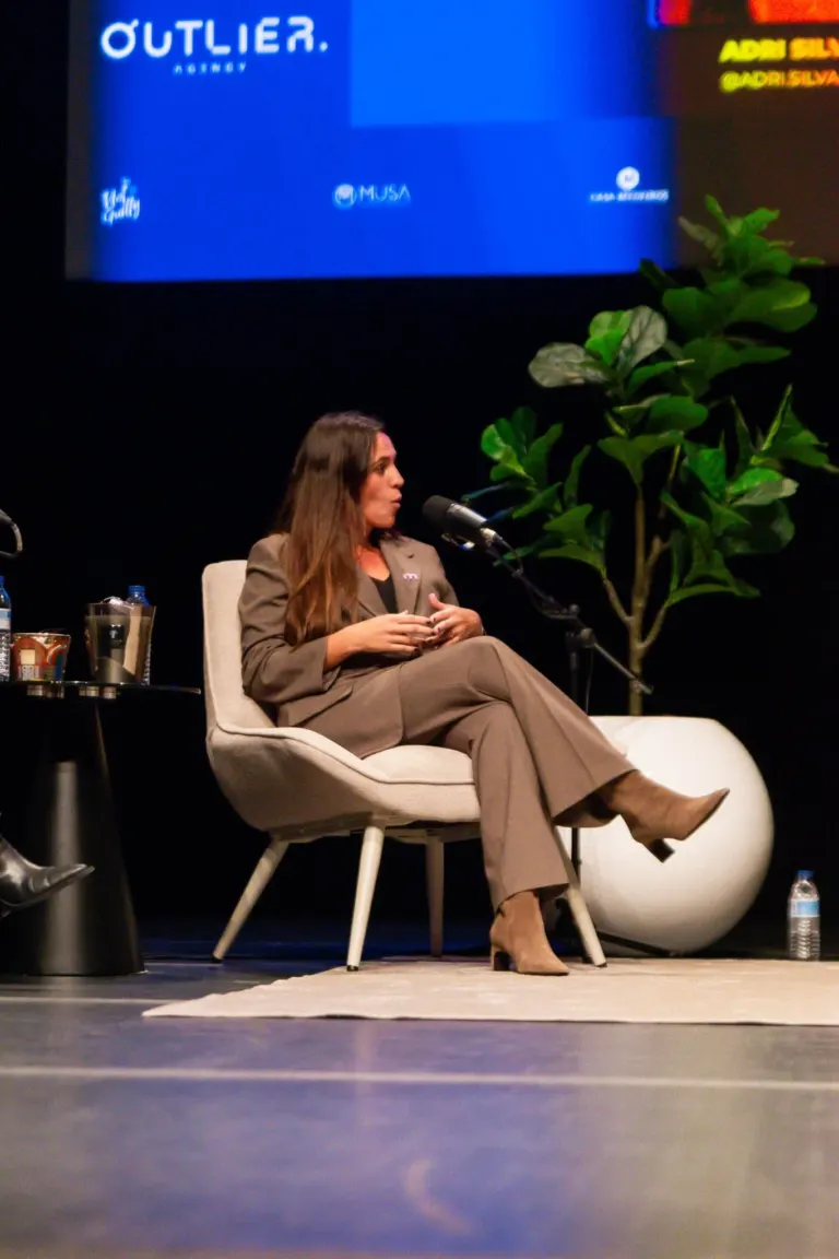 Mulher sentada em cadeira, falando em microfone, com planta ao seu lado, em um palco durante um evento.